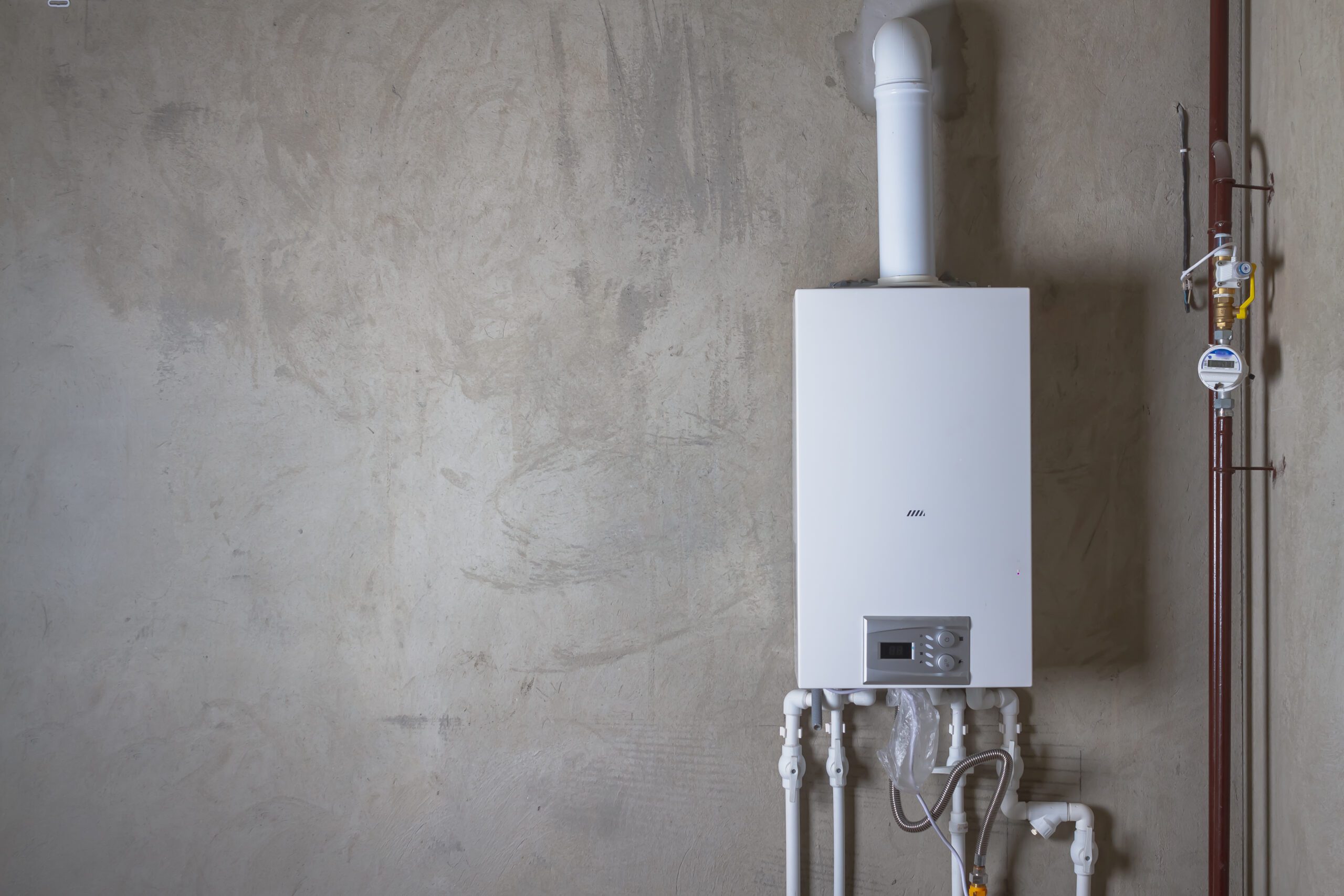 A white tankless water heater, with integrated technology and precision controls, is mounted on a concrete wall and connected to a series of pipes including a white PVC ventilation flue extending upwards and several pipes beneath, alongside an brown pipe with a gauge and a yellow lever valve.